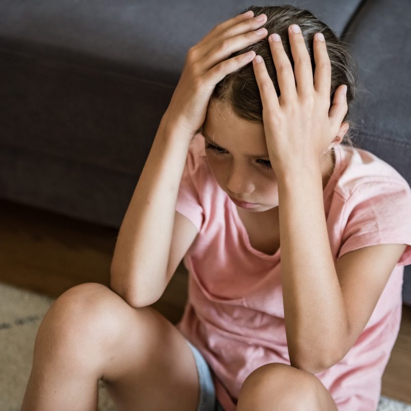 Unhappy teen girl holding her head in despair, feeling depressed, sitting on floor at home. Adolescent going through difficult times, having mental problems or mood disorder